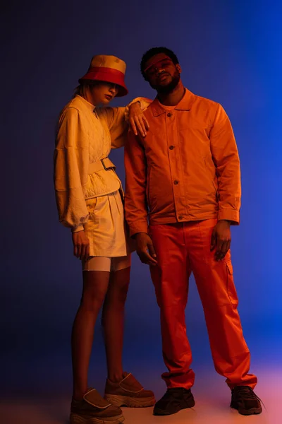 Pareja interracial elegante posando en look futurista en azul en luz naranja - foto de stock