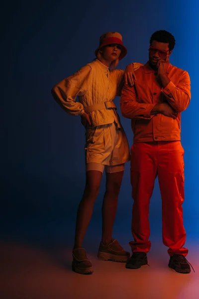 Elegante pareja multiétnica posando en look futurista sobre azul en luz naranja - foto de stock