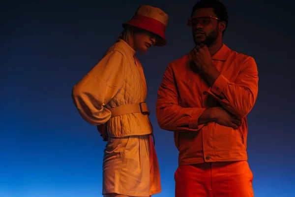 Coppia multiculturale alla moda in look futuristico in posa sul blu in luce arancione — Foto stock
