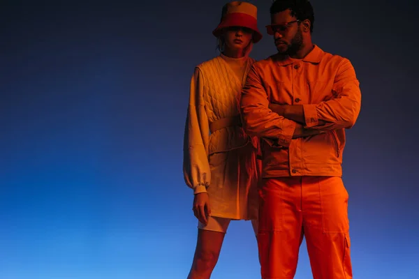 Стильна міжрасова пара в футуристичному одязі, сонцезахисних окулярах і капелюсі позує на синьому в помаранчевому світлі — стокове фото