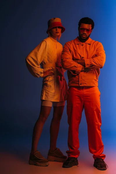 Couple interracial à la mode posant dans un look futuriste sur le bleu dans la lumière orange — Photo de stock