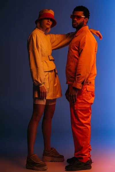 Moda casal multicultural em roupas futuristas, óculos de sol e chapéu posando em azul em luz laranja — Fotografia de Stock