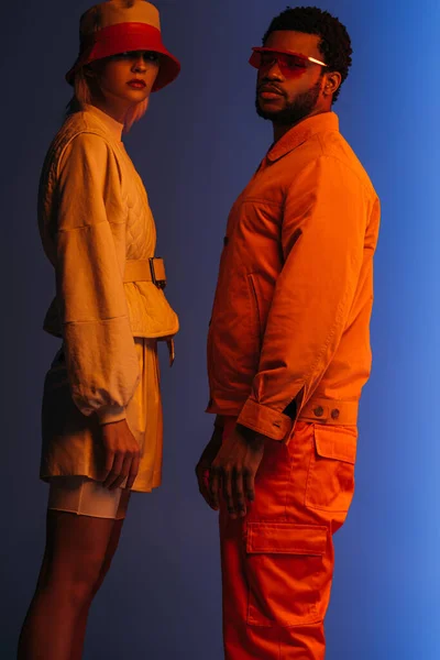 Moda casal multiétnico em roupas futuristas, óculos de sol e chapéu posando em azul em luz laranja — Fotografia de Stock