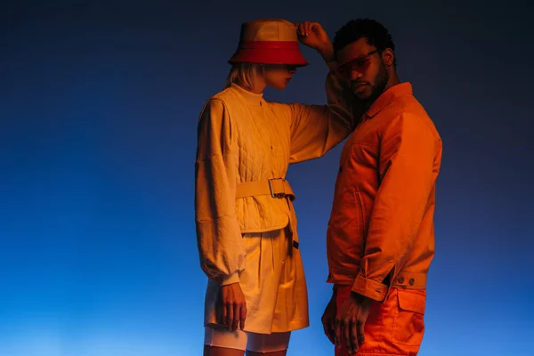 Coppia interrazziale alla moda in abiti futuristici, occhiali da sole e cappello in posa su blu in luce arancione — Foto stock