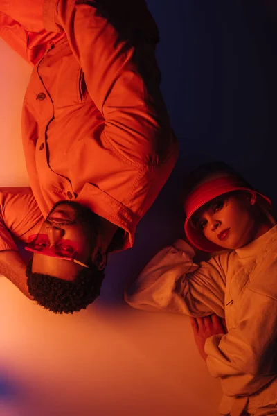 Vue de dessus du couple interracial à la mode en vêtements futuristes couché dans la lumière orange — Photo de stock