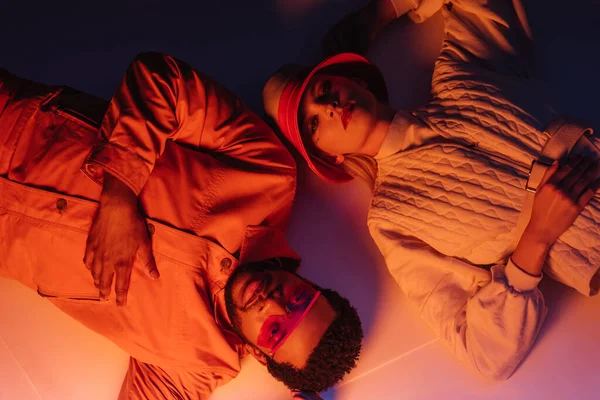 Vista superior do casal multicultural na moda em roupas futuristas deitado em luz laranja — Fotografia de Stock