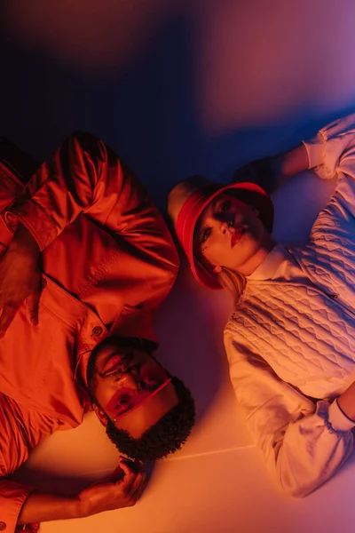 Top view of interracial couple in futuristic sunglasses and hat lying in orange light — Stock Photo