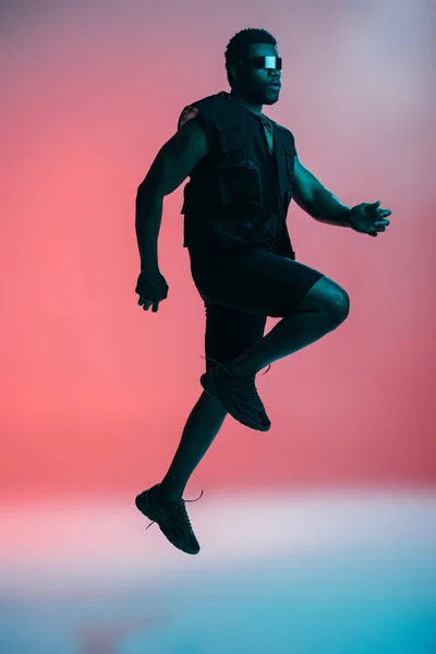 Bel homme afro-américain à la mode dans des lunettes de soleil futuristes posant sur rose à la lumière bleue — Photo de stock