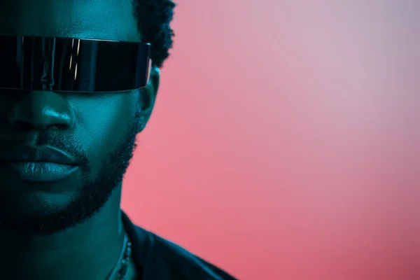 Hombre afroamericano con estilo en gafas de sol futuristas posando en rosa en luz azul - foto de stock