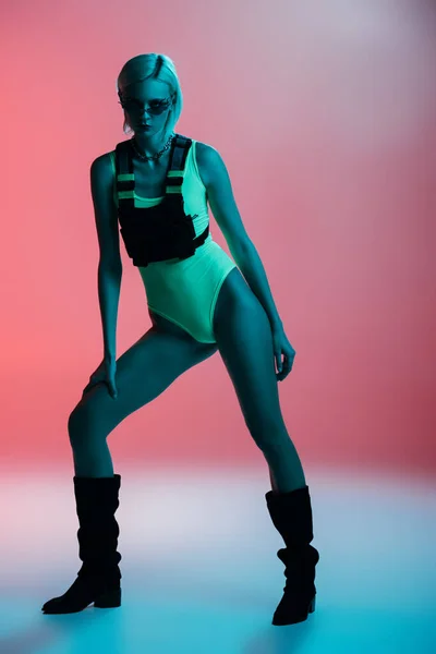 Jeune femme à la mode en body futuriste et lunettes de soleil en forme de feu posant sur rose à la lumière bleue — Photo de stock