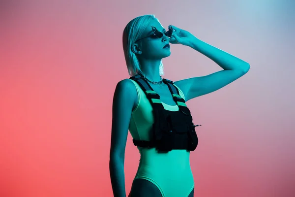 Modelo elegante em bodysuit futurista e óculos de sol em forma de fogo posando em rosa em luz azul — Fotografia de Stock