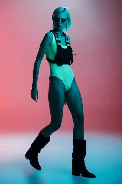 Attraktives modisches Model in futuristischem Body und feuerförmiger Sonnenbrille posiert auf rosa in blauem Licht — Stockfoto