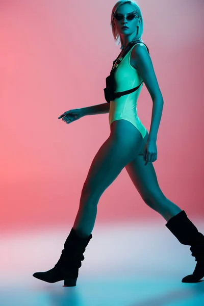 Fashionable young model in futuristic leotard and fire-shaped sunglasses posing on pink in blue light — Stock Photo
