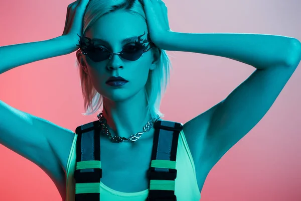 Модна блондинка-модель в футуристичному костюмі і сонцезахисних окулярах у формі вогню позує на рожевому синьому світлі — стокове фото