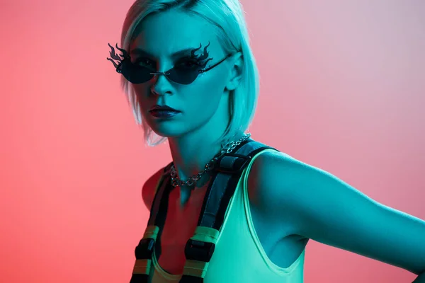 Fashionable young woman in futuristic bodysuit and fire-shaped sunglasses posing on pink in blue light — Stock Photo