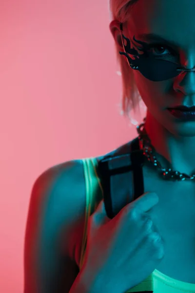 Stylisches Model in futuristischem Trikot und feuerförmiger Sonnenbrille posiert isoliert auf rosa in blauem Licht — Stockfoto