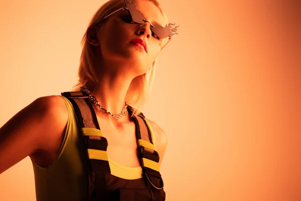 Attrayant futuriste fille posant dans des lunettes de soleil en forme de feu sur orange — Photo de stock