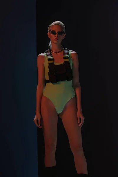 Fashionable woman in futuristic bodysuit and fire-shaped sunglasses posing on dark — Stock Photo