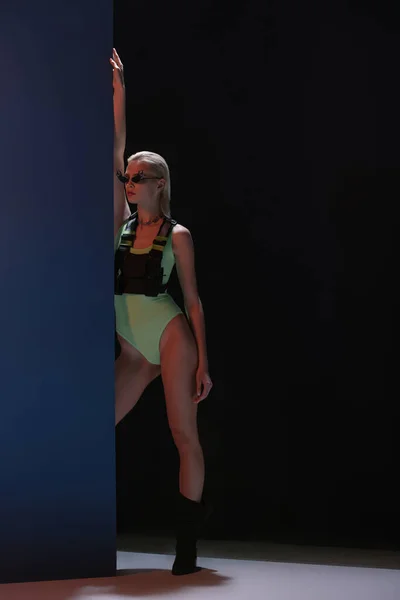 Atractiva chica de moda en traje de cuerpo futurista y gafas de sol en forma de fuego posando en la oscuridad - foto de stock