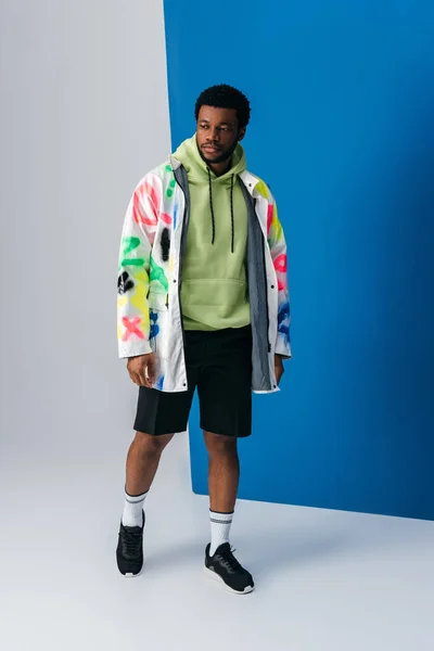 Bel homme afro-américain posant en vêtements futuristes colorés sur gris et bleu — Photo de stock