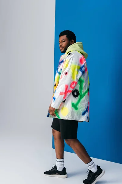 Homme afro-américain élégant posant dans des vêtements futuristes colorés sur gris et bleu — Photo de stock