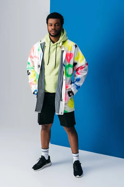 Homme afro-américain à la mode posant dans des vêtements futuristes colorés sur gris et bleu — Photo de stock