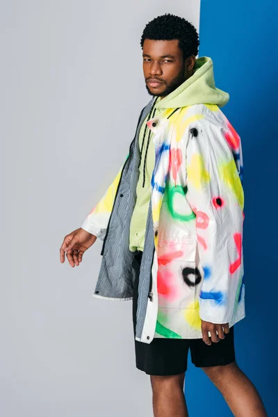 Jeune homme afro-américain à la mode posant en vêtements futuristes colorés sur gris et bleu — Photo de stock