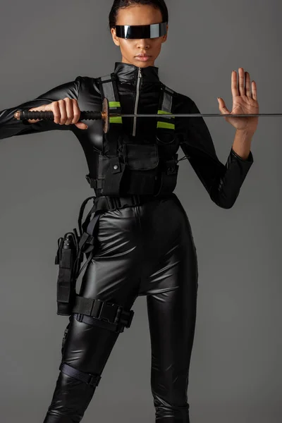 Atractiva mujer afroamericana futurista en gafas con espada aislada en gris - foto de stock