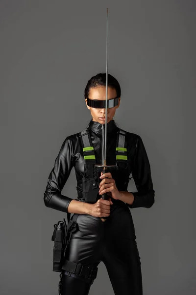 Atractiva mujer afroamericana futurista en gafas con espada aislada en gris - foto de stock