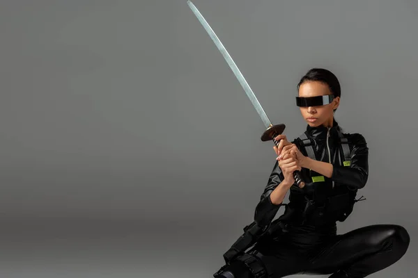 Attraktive, futuristische afrikanisch-amerikanische Frau in Brille sitzt mit Schwert auf grau — Stockfoto