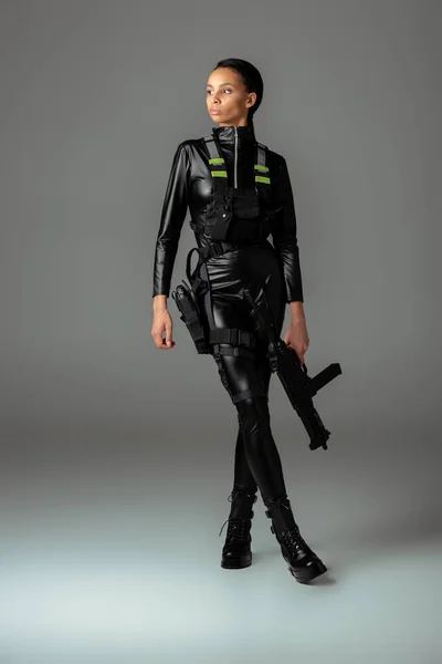 Futuristische afrikanisch-amerikanische Frau mit Sturmgewehr und gekreuzten Beinen auf grau — Stockfoto