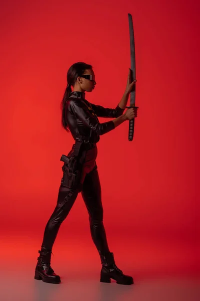 Vista lateral de mujer afroamericana futurista en gafas con espada sobre fondo rojo - foto de stock