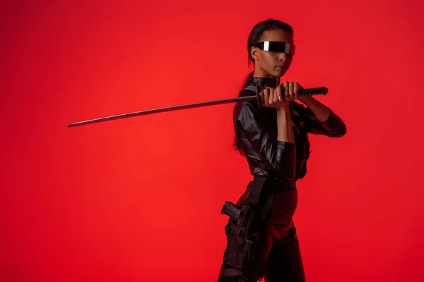 Atractiva mujer afroamericana futurista en gafas con espada aislada en rojo - foto de stock