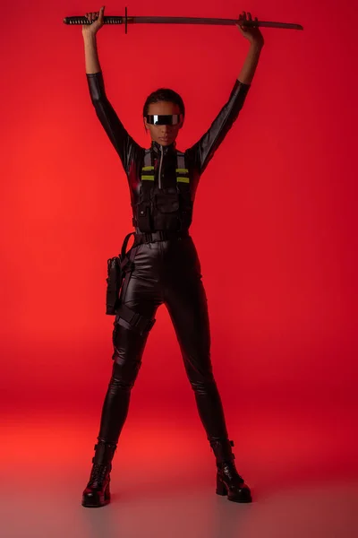 Attraktive futuristische afrikanisch-amerikanische Frau in Brille mit Schwert über dem Kopf auf rotem Hintergrund — Stockfoto