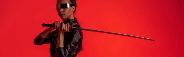Attraktive, futuristische afrikanisch-amerikanische Frau in Brille mit Schwert isoliert auf rotem, panoramischem Foto — Stockfoto