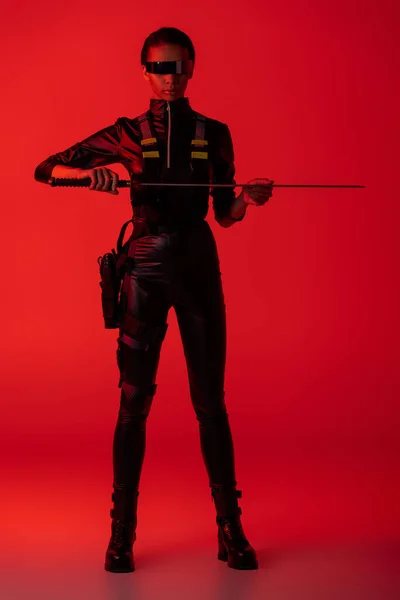 Atractiva mujer afroamericana futurista en gafas con espada sobre fondo rojo - foto de stock