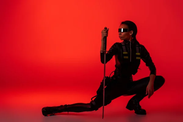 Futurista afroamericana mujer en gafas sentado con espada sobre fondo rojo - foto de stock