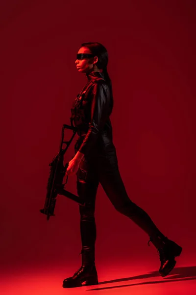 Futurista afroamericana mujer en gafas caminando con rifle de asalto sobre fondo rojo - foto de stock