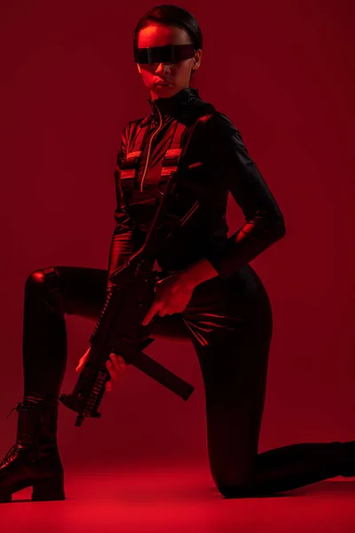 Futuristic african american woman in glasses standing on knee with assault rifle on red background — Stock Photo