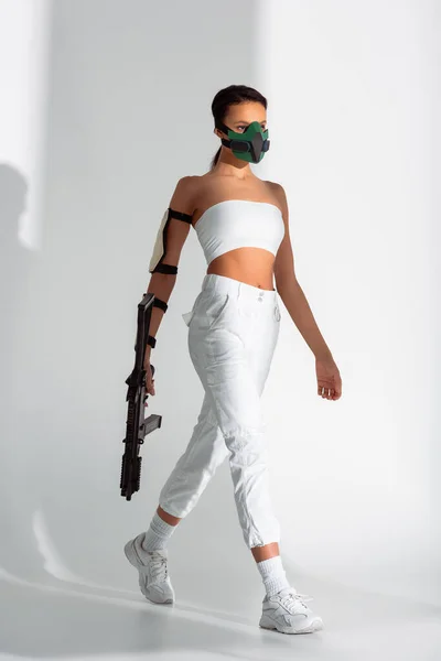Futurista afro-americana em máscara de segurança andando com rifle de assalto no fundo branco — Fotografia de Stock