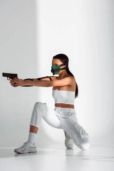 Futurista mulher afro-americana em máscara de segurança apontando arma no fundo branco — Fotografia de Stock