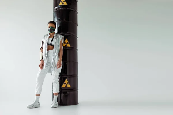 Futuristic african american woman in safety mask near radioactive waste barrels on white background — Stock Photo