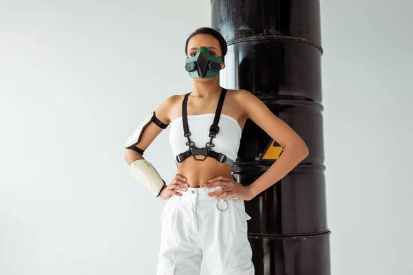 Futuristic african american woman in safety mask with hands on hips near radioactive waste barrels isolated on white — Stock Photo