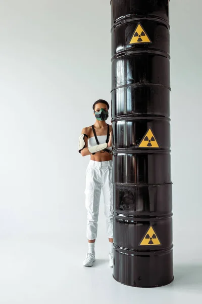 Futuristic african american woman in safety mask near radioactive waste barrels on white background — Stock Photo