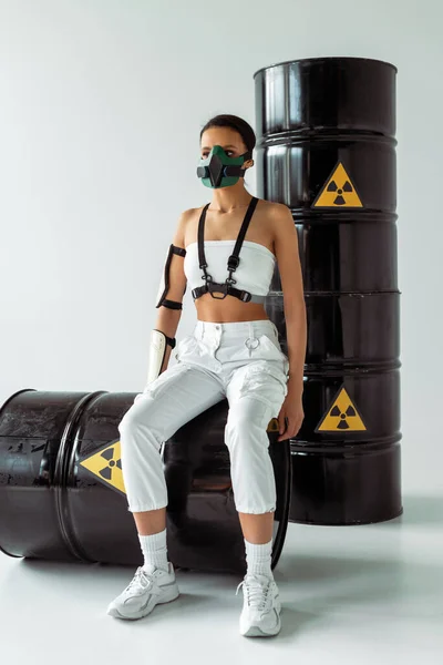 Futurista mujer afroamericana en máscara de seguridad y barriles de residuos radiactivos sobre fondo blanco — Stock Photo