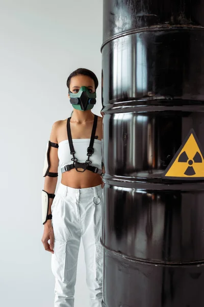 Futuristica donna afroamericana in maschera di sicurezza vicino a barili di rifiuti radioattivi isolati su bianco — Foto stock