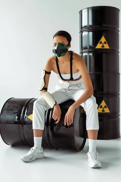 Futuristica donna afroamericana in maschera di sicurezza con pistola vicino barili di rifiuti radioattivi su sfondo bianco — Foto stock