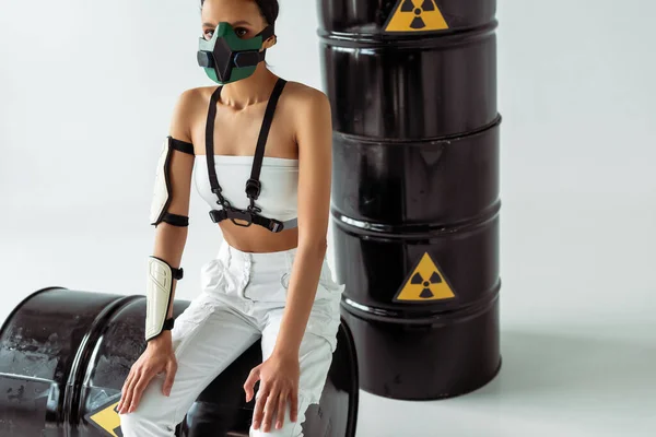Futurista mujer afroamericana en máscara de seguridad cerca de barriles de residuos radiactivos sobre fondo blanco — Stock Photo