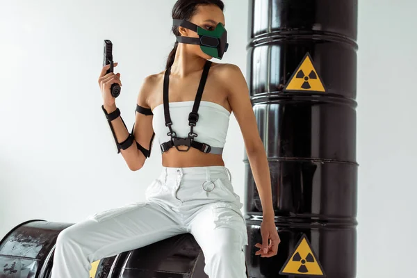 Futuristische afrikanisch-amerikanische Frau in Schutzmaske mit Pistole in der Nähe von Atommüllfässern auf weißem Hintergrund — Stockfoto