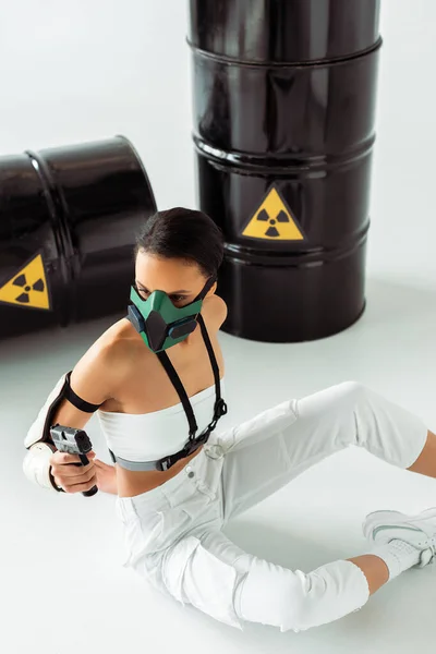 Futuristica donna afroamericana in maschera di sicurezza con pistola vicino barili di rifiuti radioattivi su sfondo bianco — Foto stock